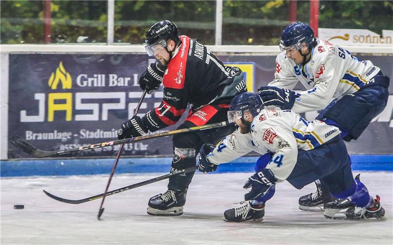 Drei Eishockeyspieler in Aktion auf dem Eis, zwei in weißen Trikots und einer in schwarzem Trikot mit rotem Streifen, alle mit Helm und Schlittschuhen.