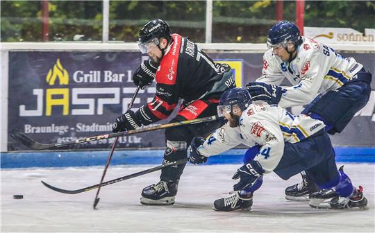 Drei Eishockeyspieler in Aktion auf dem Eis, zwei in weißen Trikots und einer in schwarzem Trikot mit rotem Streifen, alle mit Helm und Schlittschuhen.