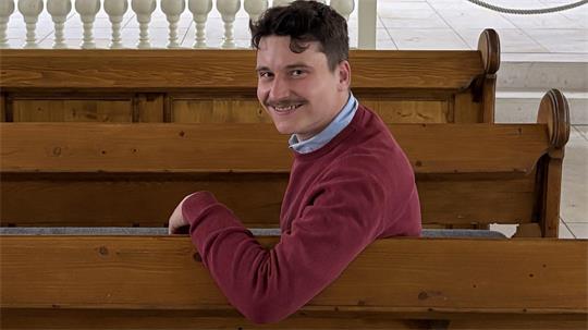 Mann mit Schnurrbart und rotem Pullover sitzt auf einer Kirchenbank und schaut lächelnd über die Schulter