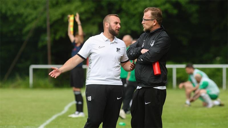 Zwei Männer in Sportkleidung unterhalten sich am Spielfeldrand eines Fußballplatzes, im Hintergrund ein Schiedsrichter mit Fahne und ein Spieler in grüner Kleidung kniet auf dem Rasen