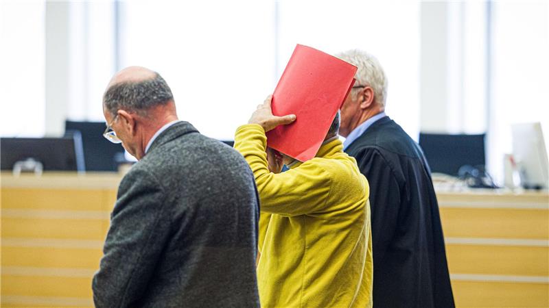 Drei Männer in einem Gerichtssaal, einer hält eine rote Mappe vor sein Gesicht, ein anderer trägt eine schwarze Robe.