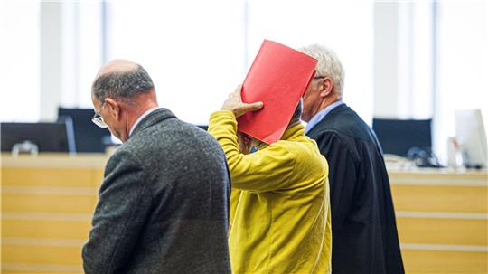 Drei Männer in einem Gerichtssaal, einer hält eine rote Mappe vor sein Gesicht, ein anderer trägt eine schwarze Robe.