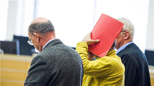 Drei Männer in einem Gerichtssaal, einer hält eine rote Mappe vor sein Gesicht, ein anderer trägt eine schwarze Robe.