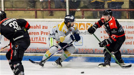 Drei Eishockeyspieler in Aktion auf dem Eis, zwei in schwarzen Trikots und einer in weißem Trikot, alle mit Helm und Schläger