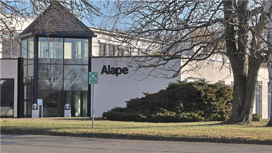 Flaches, weißes Gebäude mit großen Fenstern und dem Schriftzug 'Alape' an der Fassade, davor Bäume und Rasen.