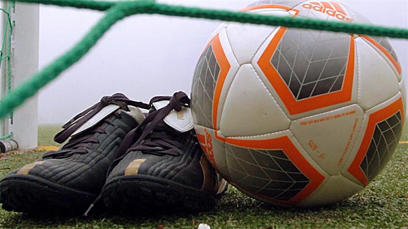 Fußballschuhe und ein Adidas-Fußball liegen auf dem Rasen hinter dem grünen Tornetz
