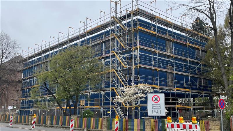 Mehrstöckiges Gebäude mit Gerüst und blauen Schutznetzen umgeben, davor Verkehrsschilder und Baustellenabsperrungen