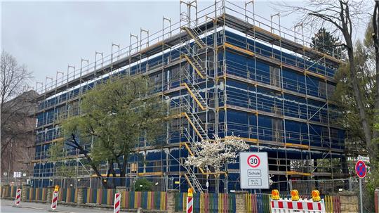Mehrstöckiges Gebäude mit Gerüst und blauen Schutznetzen umgeben, davor Verkehrsschilder und Baustellenabsperrungen