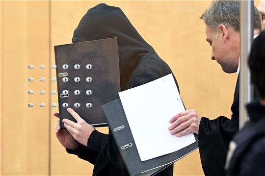 Der Angeklagte (l) verdeckt mit einem Aktenordner sein Gesicht, als er mit seinem Verteidiger zu Beginn der Verhandlung in einem Saal am Oberlandesgericht eintrifft.