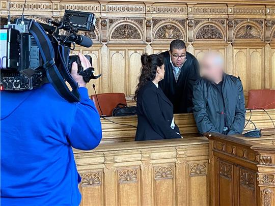 Der Angeklagte (r) steht vor Beginn des Prozesses mit seinem Anwalt Temba Hoch (hinten) und einer Dolmetscherin im Gerichtssaal im Landgericht Bremen.