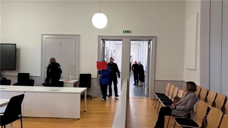 Warteraum mit Holzfußboden, Stuhlreihe rechts, mehreren Personen und offenem Durchgang zu einem Flur.