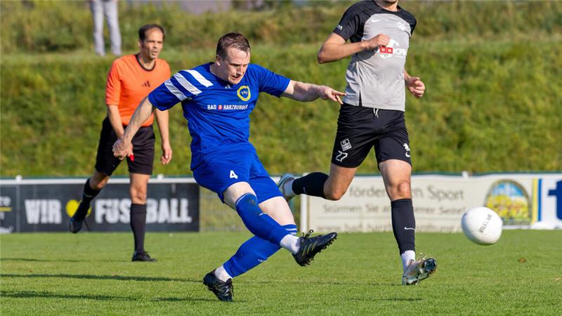 Zwei Fußballspieler in Aktion auf dem Spielfeld, einer in blauem Trikot schießt den Ball, der andere in schwarz-weißem Trikot läuft neben ihm