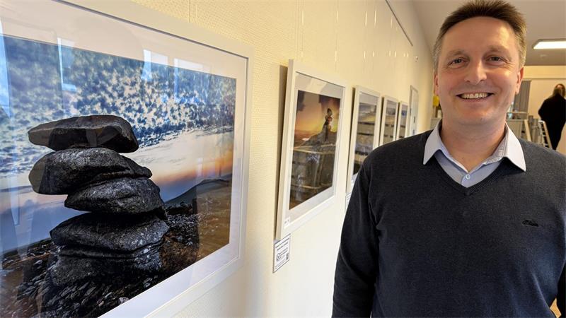 Das Foto zeigt einen Mann neben einer Wand mit Fotos.