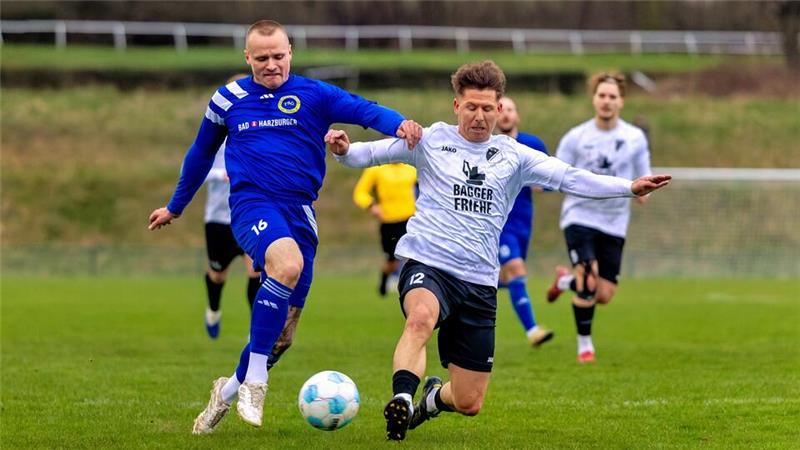 Zwei Fußballspieler in Aktion auf dem Spielfeld, einer in blauem Trikot und einer in weißem Trikot, beide kämpfen um den Ball