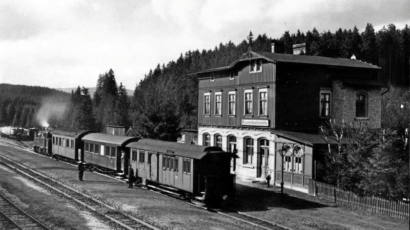 Das alte Schwarz-Weiß-Foto zeigt eine Dampflok mit drei Waggons in einem Bahnhof.