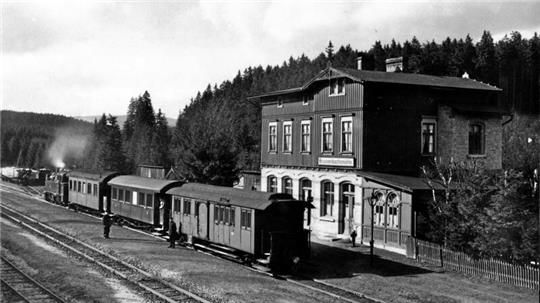 Das alte Schwarz-Weiß-Foto zeigt eine Dampflok mit drei Waggons in einem Bahnhof.
