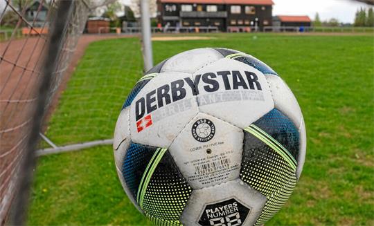 Fußball mit der Aufschrift 'DERBYSTAR' im Tornetz auf einem Fußballfeld mit Gebäuden im Hintergrund