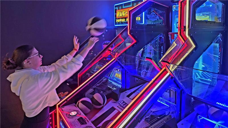 Person mit Zopf und Kapuzenpullover spielt an einem beleuchteten Arcade-Spielautomaten in einer dunklen Spielhalle.