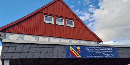 Fassade eines roten Hauses mit drei kleinen Fenstern im Giebel und einem blauen Schild mit weißer Schrift und gelbem Logo an der unteren Wand