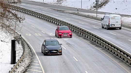 Zwei Autos fahren auf einer mehrspurigen, leicht verschneiten Straße, ein schwarzes Auto in der linken Spur und ein rotes in der mittleren Spur, rechts eine weiße Transporter auf der rechten Spur.