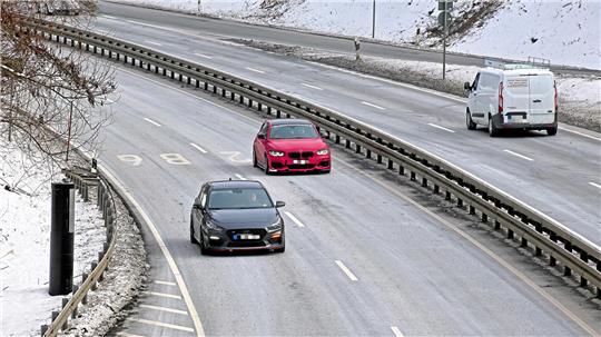 Zwei Autos fahren auf einer mehrspurigen, leicht verschneiten Straße, ein schwarzes Auto in der linken Spur und ein rotes in der mittleren Spur, rechts eine weiße Transporter auf der rechten Spur.