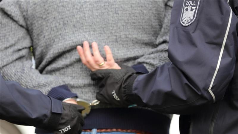 Der Rücken eines Mannes ist zusehen. Zwei Polizisten halten seine Hände an seinem unteren Rücken fest.