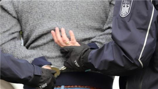 Der Rücken eines Mannes ist zusehen. Zwei Polizisten halten seine Hände an seinem unteren Rücken fest.