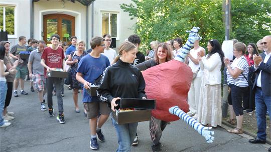 Gruppe von Jugendlichen trägt eine große, rot-blaue Skulptur mit zwei langen Stangen, umgeben von applaudierenden Erwachsenen vor einem Gebäude