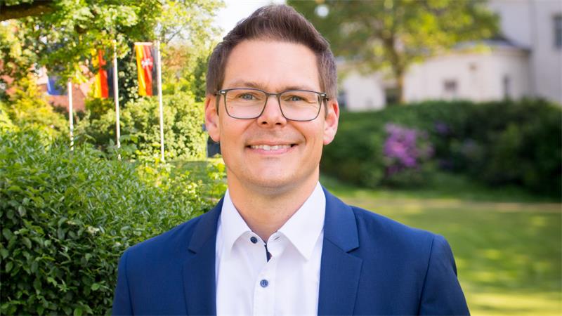 Mann mit Brille und blauem Sakko lächelt vor grüner Gartenlandschaft mit Fahnen im Hintergrund