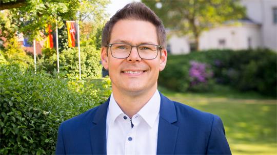 Mann mit Brille und blauem Sakko lächelt vor grüner Gartenlandschaft mit Fahnen im Hintergrund