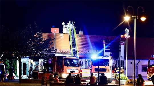 Zwei Einsatzfahrzeuge der Feuerwehr stehten vor einem Gebäude. Bei einem Fahrzeug ist die Leiter ausgefahren. Vor den Einsatzfahrzeugen sitzen Menschen und beobachten die Löscharbeiten. 