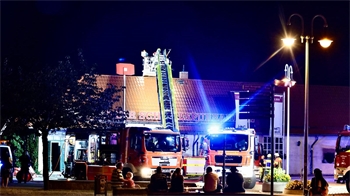 Zwei Einsatzfahrzeuge der Feuerwehr stehten vor einem Gebäude. Bei einem Fahrzeug ist die Leiter ausgefahren. Vor den Einsatzfahrzeugen sitzen Menschen und beobachten die Löscharbeiten. 