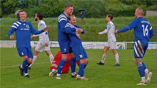 Fußballspieler in blauen und weißen Trikots auf einem Spielfeld mit grüner Wiese und Bäumen im Hintergrund.