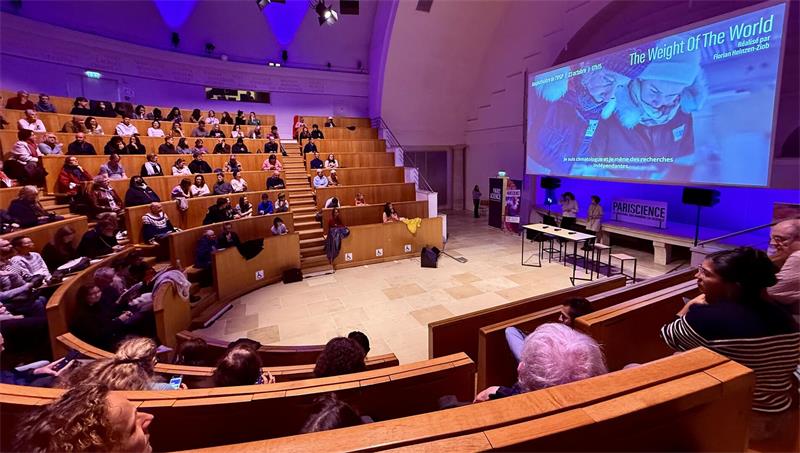 Auditorium mit Zuschauern auf hölzernen Sitzreihen und einer großen Leinwand mit der Überschrift 'The Weight Of The World'
