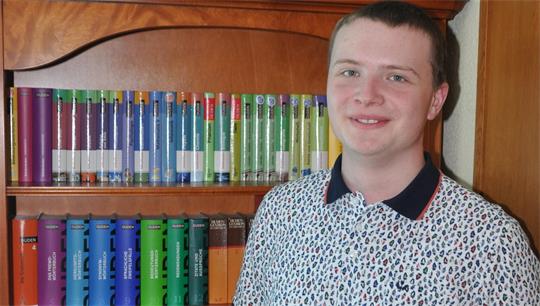 Junger Mann mit kurz geschnittenem Haar und gemustertem Hemd steht vor einem Bücherregal mit bunten Büchern