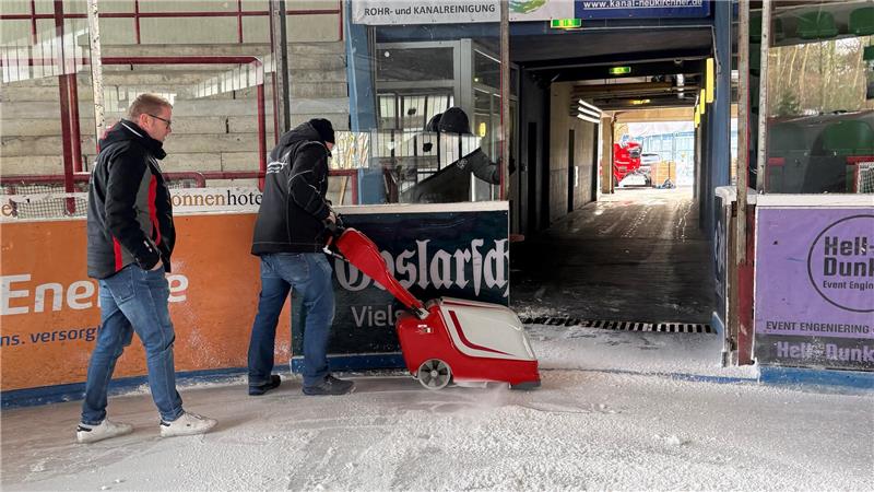 Ein Mann schiebt eine Art Rasenmäher unter den Anweisungen eines anderen Mannes an der Bande des Eisstadions entlang.