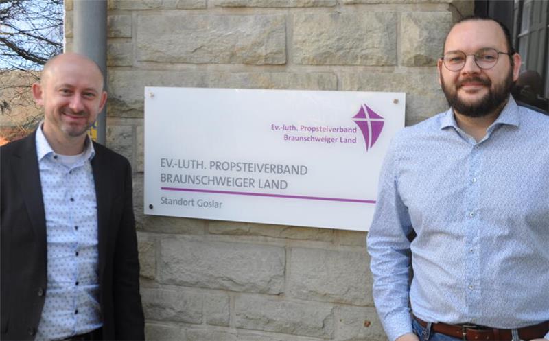 Zwei Männer stehen neben einem Schild mit der Aufschrift 'EV.-LUTH. PROPSTEIVERBAND BRAUNSCHWEIGER LAND Standort Goslar' an einer Steinwand