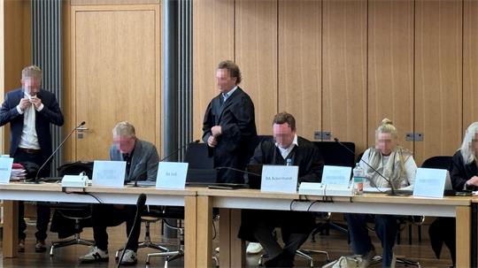 Fünf Personen sitzen an einem langen Tisch vor einer Holzpaneelwand, zwei von ihnen tragen Mund-Nasen-Schutz.