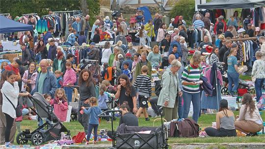Gruppe von Menschen vor mehreren Tischen mit verschiedenen Gegenständen auf einem Flohmarkt im Freien.