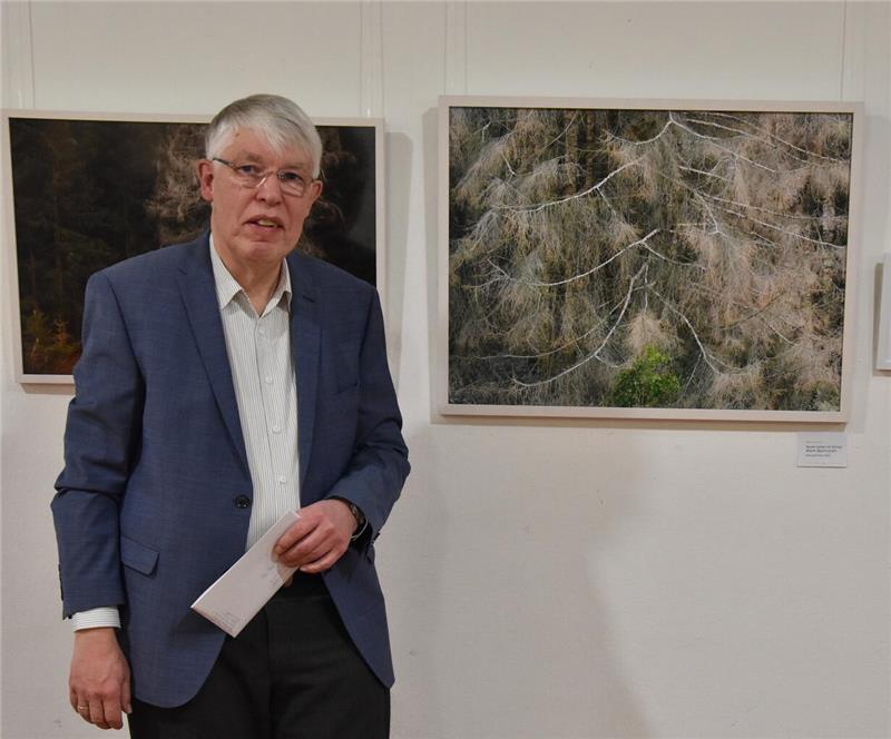 Bertram Köster in blauem Sakko und weißem Hemd steht vor gerahmten Fotografien von Bäumen an einer weißen Wand.