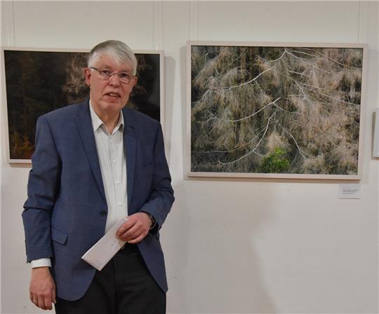 Bertram Köster in blauem Sakko und weißem Hemd steht vor gerahmten Fotografien von Bäumen an einer weißen Wand.