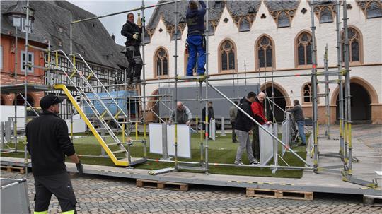 Mehrere Personen bauen ein Gerüst mit gelben Treppenstufen auf einem gepflasterten Platz vor einem historischen Gebäude mit gotischen Fenstern auf