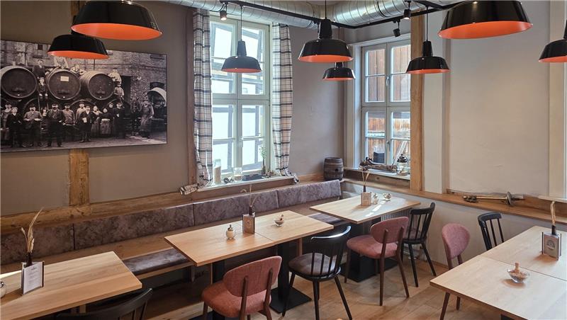 Gastraum mit Holztischen, gepolsterten Stühlen und großen Fenstern, an der Wand ein schwarz-weißes Foto und orangefarbene Hängelampen.