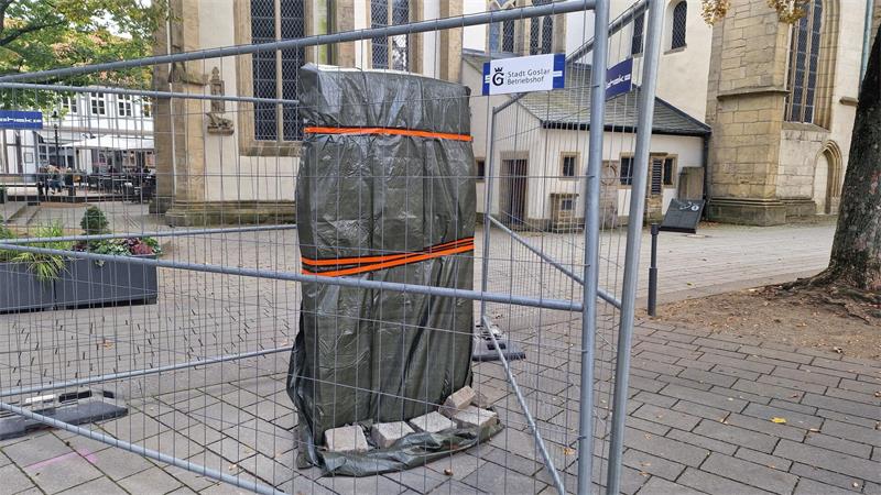 Mit Planen und Bändern gesicherte Statue hinter Bauzaun auf gepflastertem Platz vor Kirche