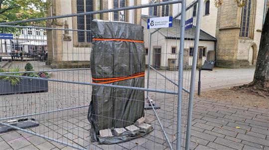 Mit Planen und Bändern gesicherte Statue hinter Bauzaun auf gepflastertem Platz vor Kirche