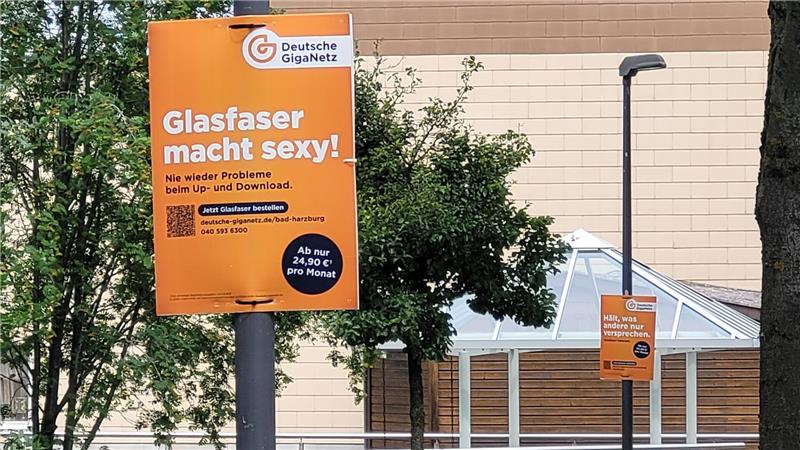 Zwei orangefarbene Werbeschilder der Deutschen GigaNetz an Straßenlaternen vor einem Gebäude und Bäumen