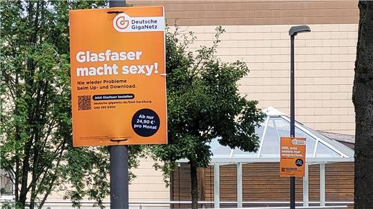 Zwei orangefarbene Werbeschilder der Deutschen GigaNetz an Straßenlaternen vor einem Gebäude und Bäumen