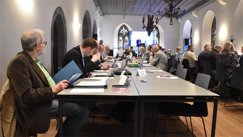 Menschen sitzen an einem langen Tisch in einem historischen Raum mit hohen Fenstern und Kronleuchtern und arbeiten an Laptops oder lesen Unterlagen