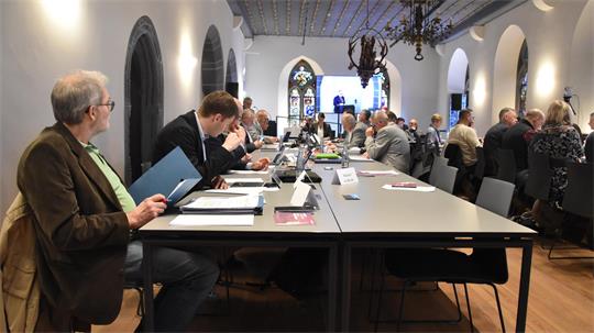 Menschen sitzen an einem langen Tisch in einem historischen Raum mit hohen Fenstern und Kronleuchtern und arbeiten an Laptops oder lesen Unterlagen