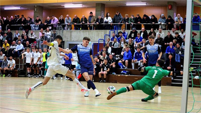 Hallenszene eines Fußballspiels mit mehreren Spielern in Aktion, ein Spieler in grün rutscht zum Ball, Zuschauer sitzen auf Tribünen im Hintergrund